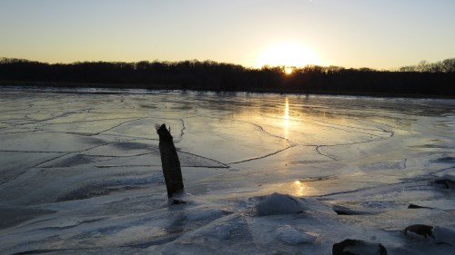 Sunset over a frozen lake, with ice patterns and a silhouette of trees in the background.