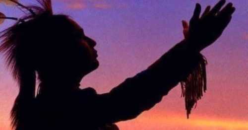 Silhouette of a person with a feathered headdress, arms raised against a colorful sunset sky.