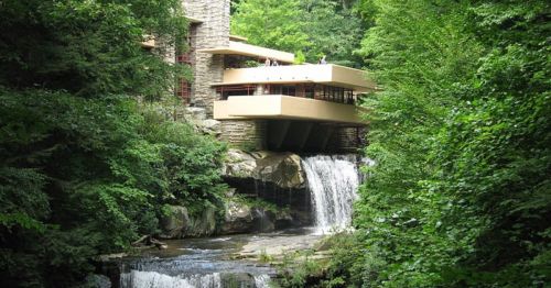 A modern building with a flat roof sits above a waterfall, surrounded by lush green trees.