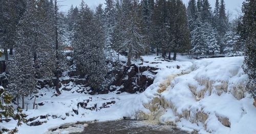 A snowy landscape featuring a waterfall surrounded by evergreen trees and a rocky shoreline.