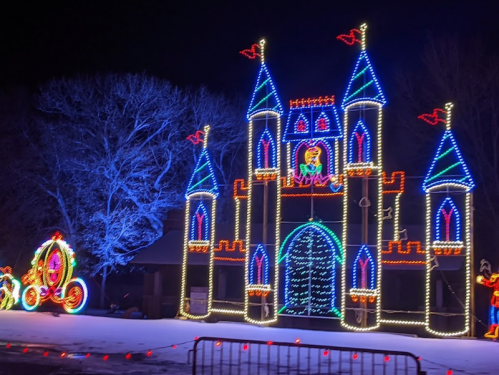 A brightly lit castle made of colorful lights, with a carriage nearby, set against a dark night sky.