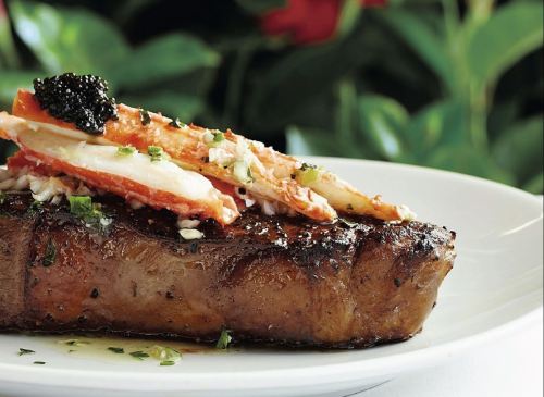 A grilled steak topped with crab meat and caviar, garnished with herbs, served on a white plate.