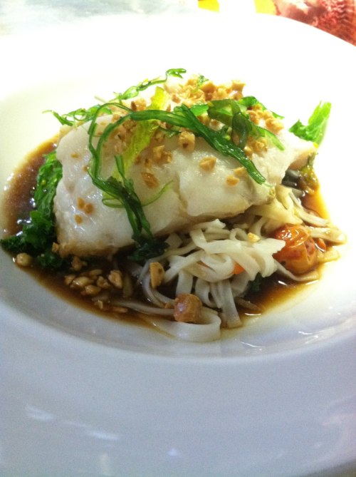 A plate of steamed fish topped with herbs and crushed peanuts, served on a bed of noodles and greens in a savory sauce.