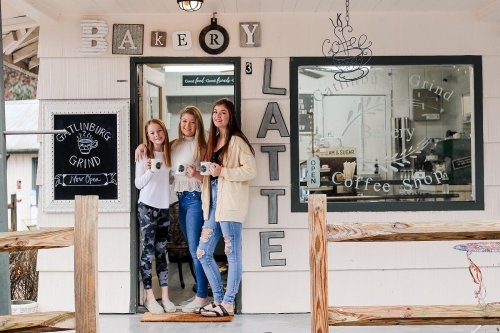 Three young women stand in the doorway of a bakery and coffee shop, smiling and holding mugs. Signs decorate the exterior.