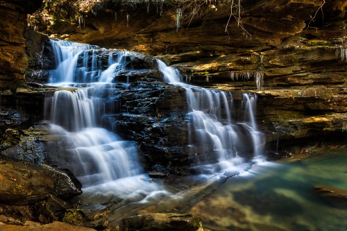 A serene waterfall cascades over rocky ledges into a clear pool, surrounded by lush greenery and natural rock formations.