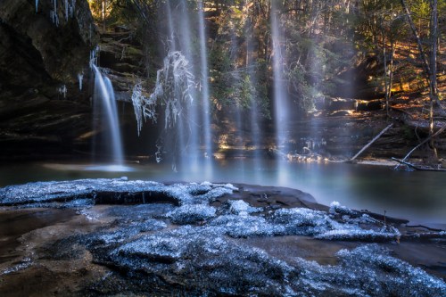 A serene waterfall cascades over icy rocks, surrounded by lush greenery and mist in a tranquil forest setting.