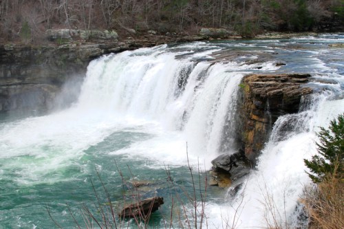 A cascading waterfall flows over rocky cliffs into a turquoise pool, surrounded by trees and rugged terrain.