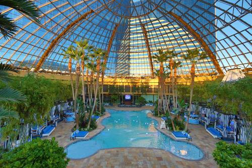 Indoor pool surrounded by palm trees and lounge chairs, under a glass dome with reflections of nearby buildings.