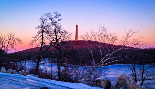 A tall monument on a hill at sunset, surrounded by bare trees and a frozen lake, with a colorful sky.