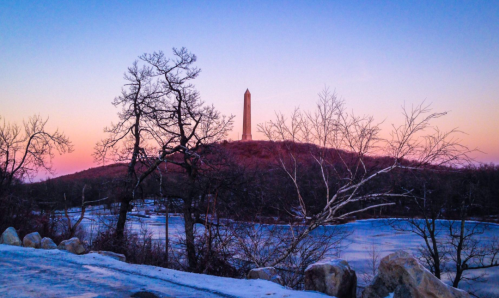 A tall monument on a hill at sunset, surrounded by bare trees and a frozen lake, with a colorful sky.