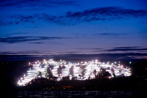 A snowy hill illuminated by bright lights at dusk, with a dark sky and clouds above.