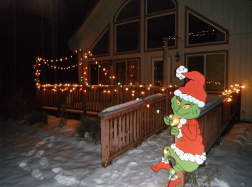 A cartoon Grinch in a Santa hat sits on a snowy porch decorated with colorful Christmas lights.