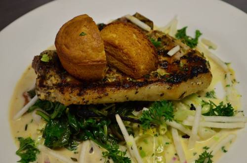 Grilled fish fillet topped with a small bread roll, served on a bed of greens and drizzled with a creamy sauce.