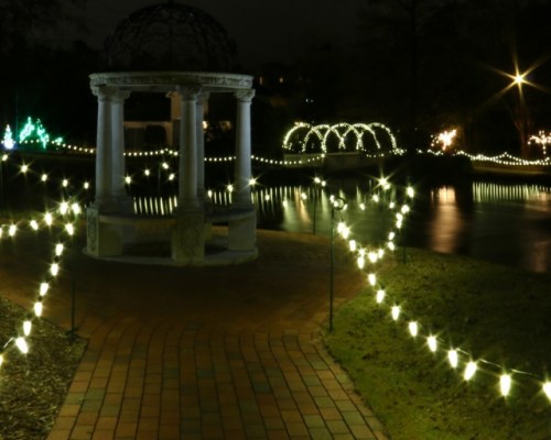 A serene night scene featuring a gazebo surrounded by glowing lights along a pathway by a calm water body.