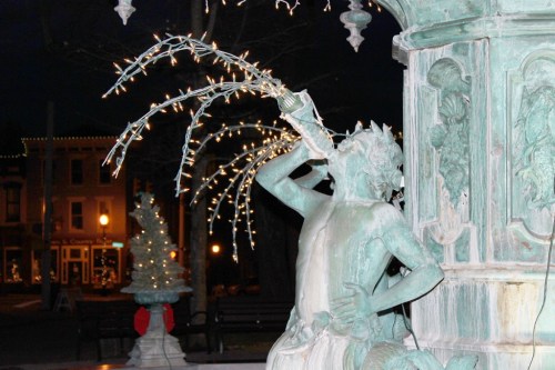 A night scene of a fountain statue blowing water, surrounded by festive lights and holiday decorations.