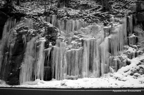 Icicles and frozen water cascading down a rocky hillside, surrounded by snow in a black and white landscape.