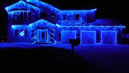 A house decorated with blue Christmas lights, glowing brightly against a dark night sky. Snow covers the ground.