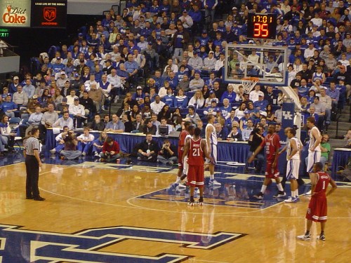 A basketball game in a packed arena, with players on the court and fans in blue cheering in the stands.