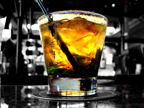 A close-up of a glass filled with a golden drink, ice, and a straw, set against a blurred background.