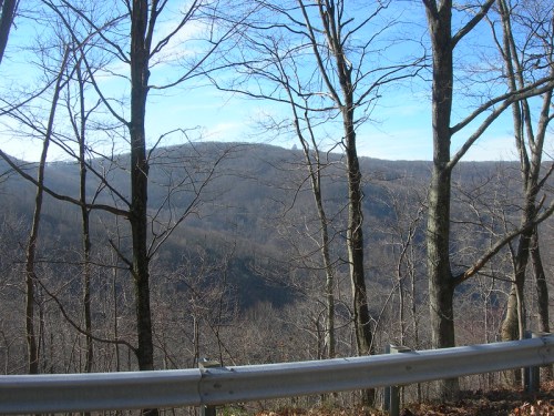 A scenic view of a mountainous landscape framed by bare trees and a guardrail under a clear blue sky.