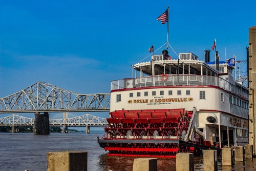 A riverboat docked by the shore, with a bridge in the background and clear blue skies above.