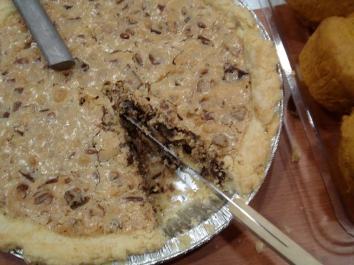 A pecan pie with a slice cut out, resting on a wooden surface next to some muffins.