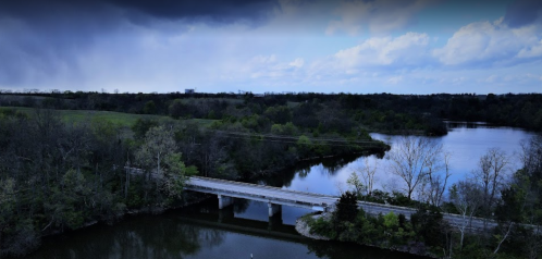 Aerial view of a river with a bridge, surrounded by lush greenery and a cloudy sky.