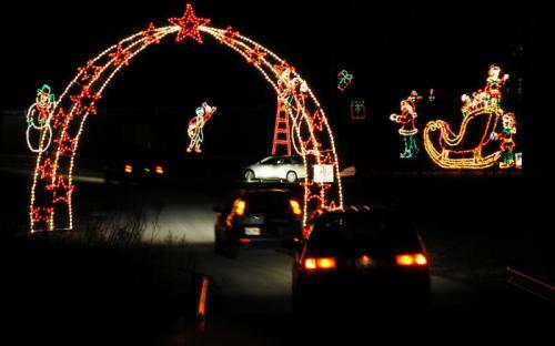 Cars drive under a festive arch of colorful holiday lights, featuring snowmen, Santa, and a sleigh in a nighttime scene.