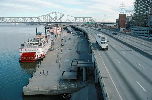 A riverfront scene featuring a steamboat, a highway, and a bridge in the background, with people walking along the dock.