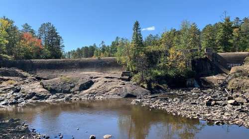 A serene landscape featuring a rocky riverbank, a dam, and trees with autumn foliage under a clear blue sky.