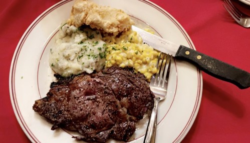 A plate with a juicy steak, fried chicken, mashed potatoes with gravy, and corn, set on a red tablecloth.