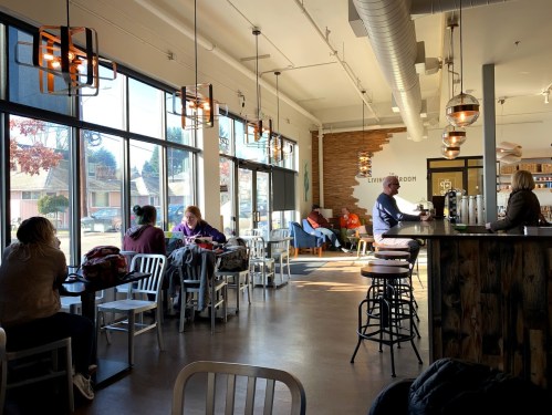A cozy café interior with large windows, patrons seated at tables, and a barista serving coffee at the counter.