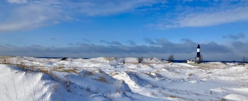 A snowy landscape with rolling dunes and a lighthouse in the distance under a blue sky with scattered clouds.