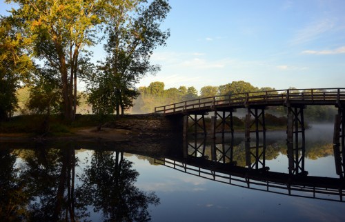 A wooden bridge spans a calm river, surrounded by trees and mist, reflecting the serene landscape in the water.