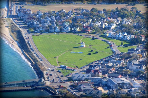 Aerial view of a coastal town with green park, houses, and a beach along the shoreline.