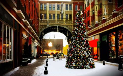 A festive alleyway with a decorated Christmas tree, snow on the ground, and shops lining the sides.
