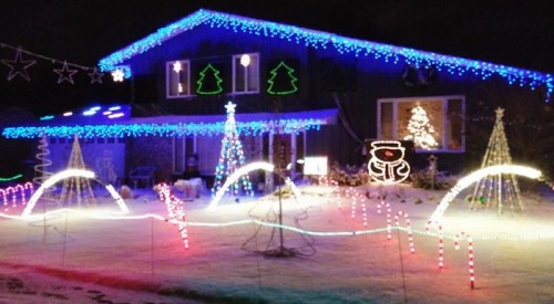 A house decorated with colorful Christmas lights, featuring trees, snowmen, and festive designs in a snowy yard.