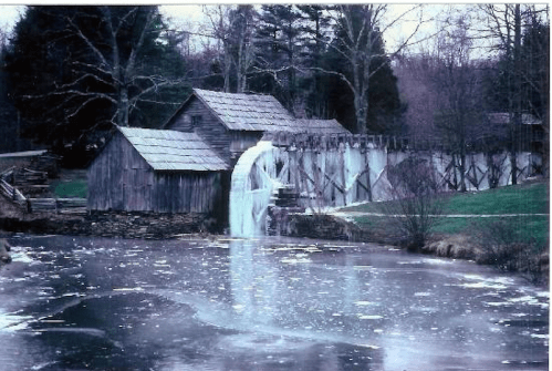 A rustic wooden mill beside a frozen pond, with a waterwheel partially covered in ice, surrounded by trees.