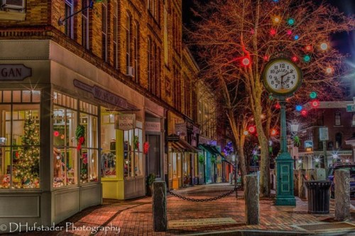 A charming street at night, adorned with festive lights and a clock, showcasing holiday decorations in shop windows.