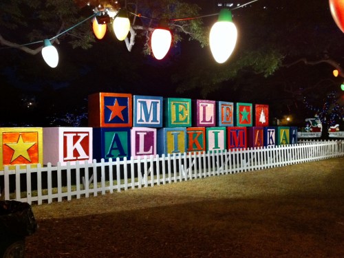 Colorful holiday blocks spell "Mele Kalikimaka" with festive lights in the background and a white picket fence.