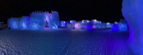 A panoramic view of an illuminated ice sculpture park at night, featuring colorful lights and snow-covered ground.