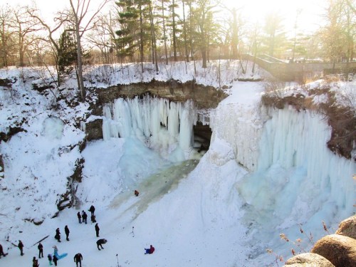 A frozen waterfall surrounded by snow and trees, with people exploring the icy landscape below.