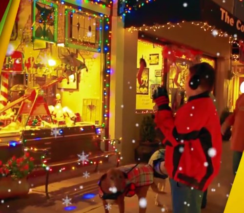 A festive street scene with holiday lights, a person waving, and a dog in a sweater amidst falling snow.