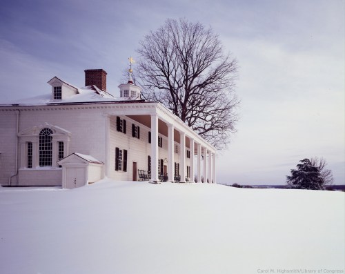 A historic white house with columns, surrounded by deep snow and a bare tree under a cloudy sky.