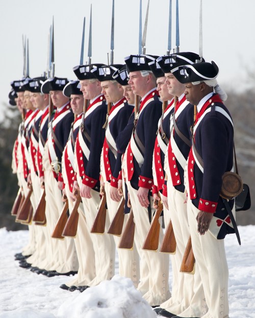 A line of soldiers in historical uniforms, standing in snow, holding rifles with bayonets, facing forward.