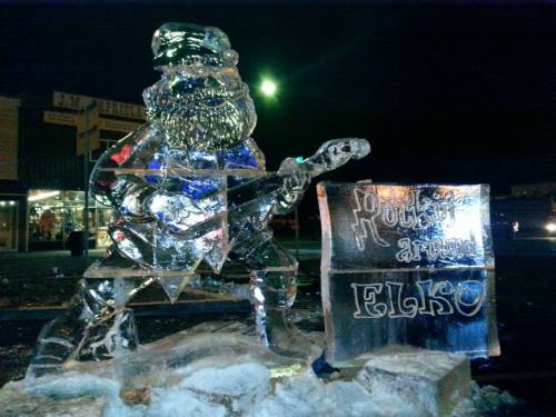 An ice sculpture of a bear playing guitar, with a sign reading "Polar Art" and "Elko" nearby, illuminated at night.
