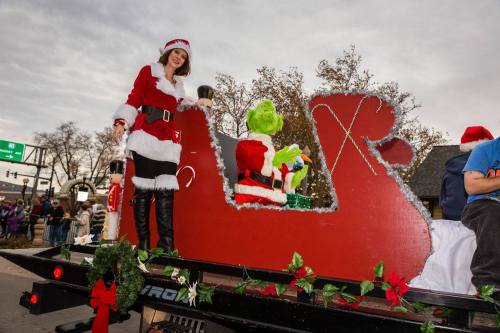 A woman in a Santa outfit stands on a festive float with the Grinch, surrounded by holiday decorations.