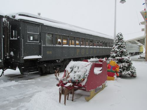 A snowy scene featuring a black train car labeled "Polar," a decorated Christmas tree, and a festive sleigh.