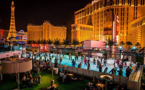 A vibrant outdoor ice skating rink at night, surrounded by illuminated buildings and palm trees in a lively city setting.