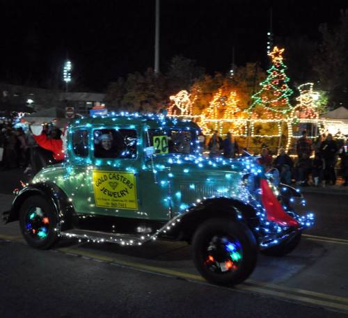 A vintage car decorated with colorful lights and a festive sign, driving through a night parade with holiday decorations.